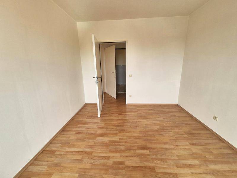 Leerer Raum mit Laminatboden und hellen Wänden, eine offene Tür führt in den nächsten Bereich.