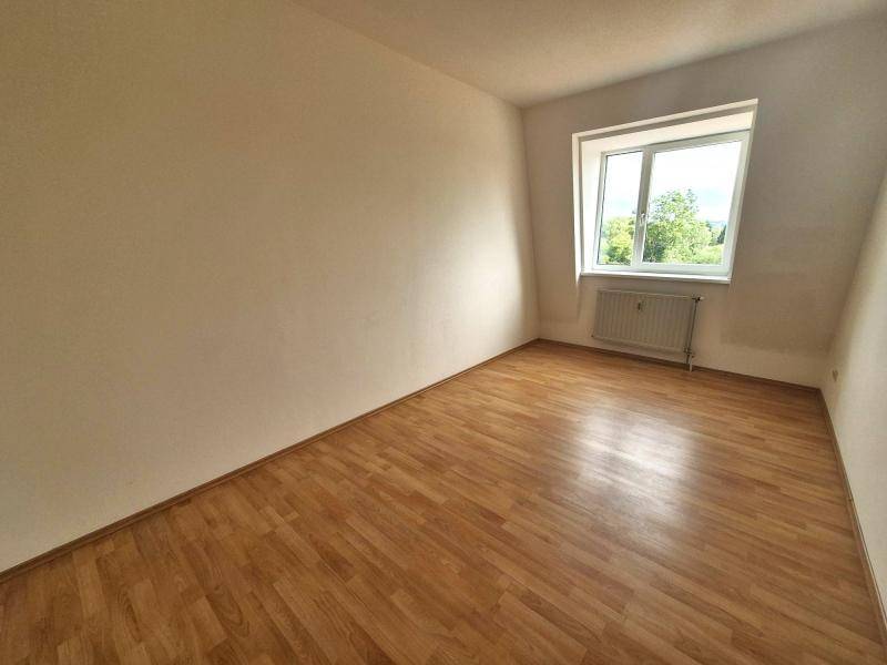 Helles Zimmer mit Dachschräge, Laminatboden und einem Fenster mit Blick ins Grüne.