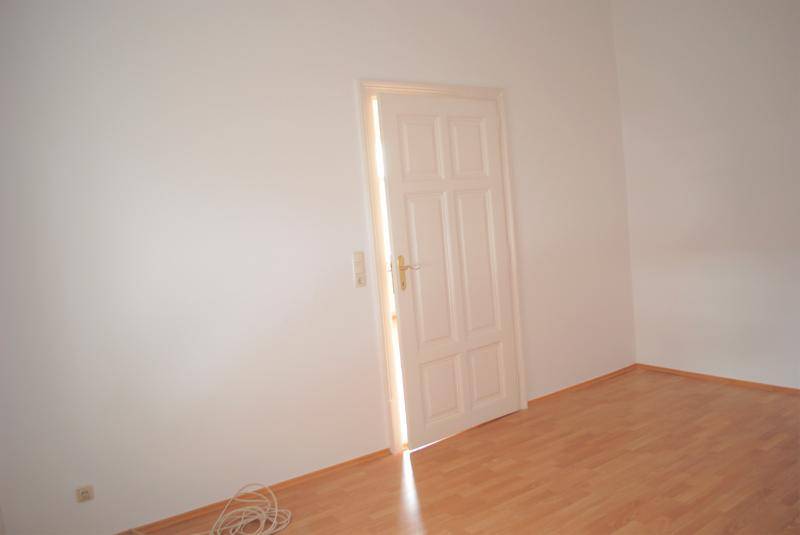 Leerer Raum mit geschlossener Holztür und hellem Laminatboden, durch die Tür fällt Sonnenlicht.
