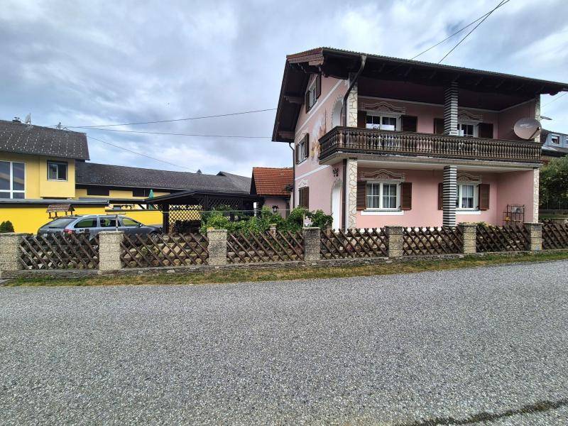 Traditionelles Haus mit rosafarbener Fassade, Holzbalkon und Lattenzaun, umgeben von Nachbargebäuden.