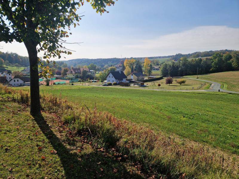 Panoramablick über grüne Felder auf ein Dorf mit Häusern und einem Tennisplatz in der Ferne.