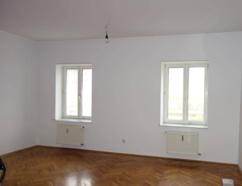 Geräumiges Zimmer mit weiß gestrichenen Wänden, zwei Fenstern und glänzendem Fischgrätparkett.