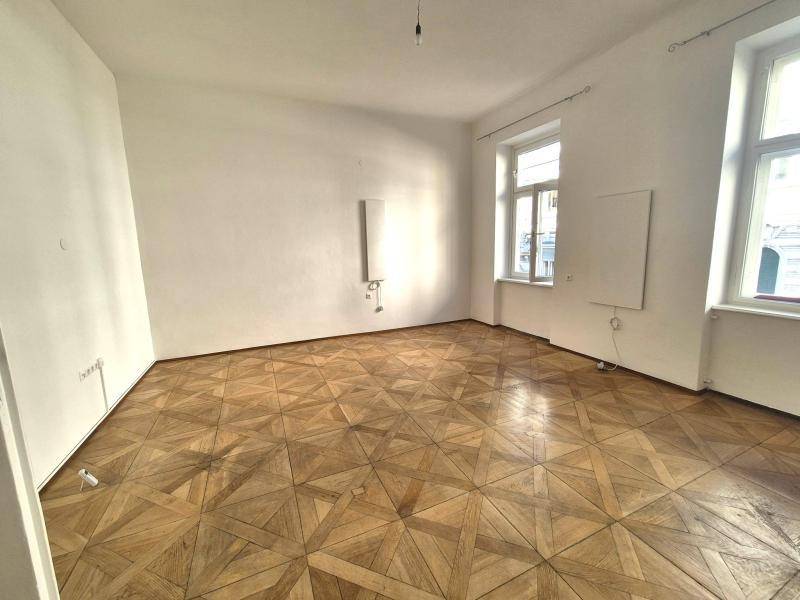 Helles Zimmer mit klassischem Parkettboden und zwei Fenstern, bereit zur individuellen Gestaltung und Nutzung.