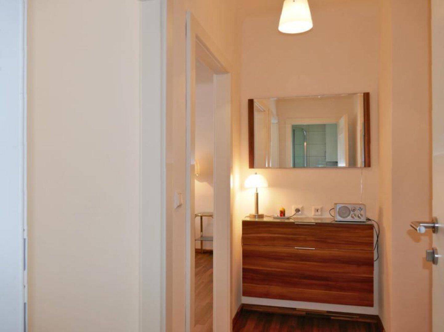 Heller Eingangsbereich mit einem Spiegel, einer Holzkommode und einer dekorativen Tischlampe.