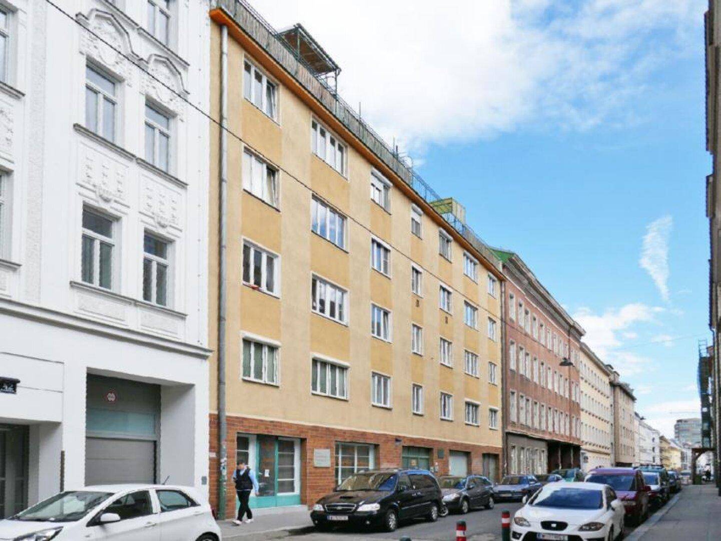 Mehrstöckiges Wohngebäude an einer Stadtstraße mit geparkten Autos und blauem Himmel.