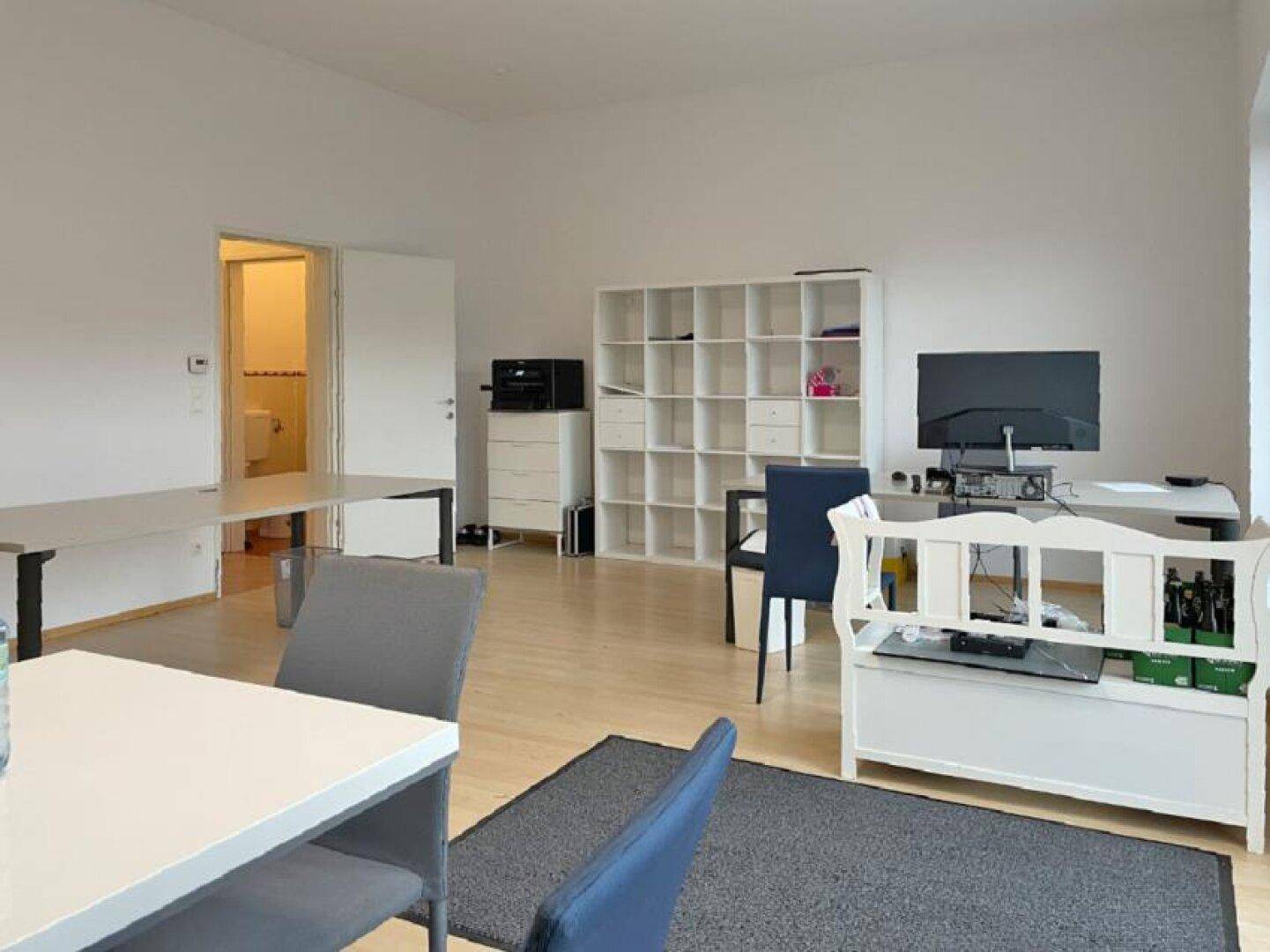 Bürofläche mit Regalen, Arbeitsplätzen und Zugang zu einem angrenzenden Sanitärbereich mit Toilette.