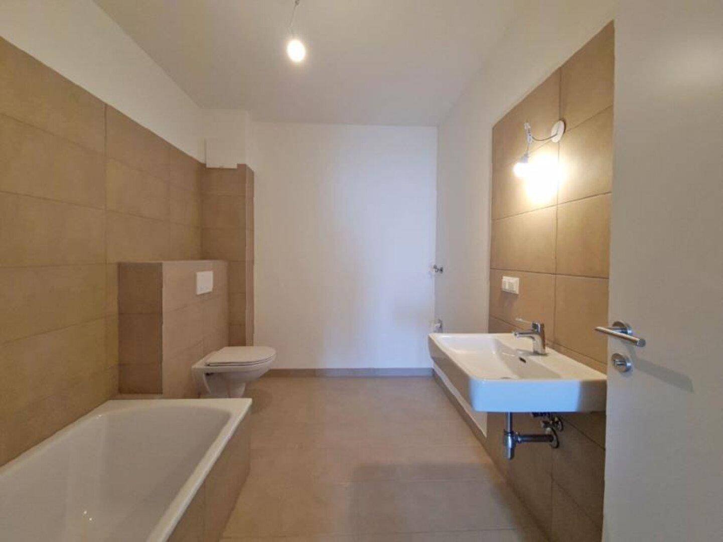 Geräumiges Badezimmer mit Badewanne, Wand-WC, modernem Waschbecken und beigefarbenen Fliesen.