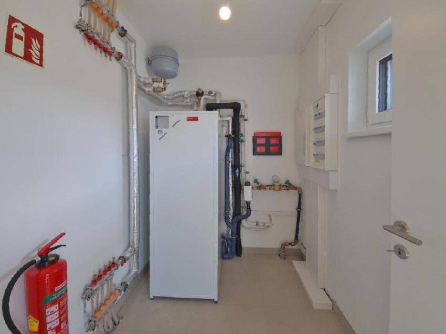 Technikraum mit Heizungsanlage, Rohrleitungen und einem Fenster für Belüftung.