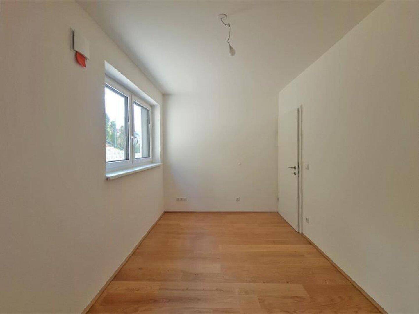 Leeres Zimmer mit Holzboden und einem Fenster, das viel Tageslicht hereinlässt.