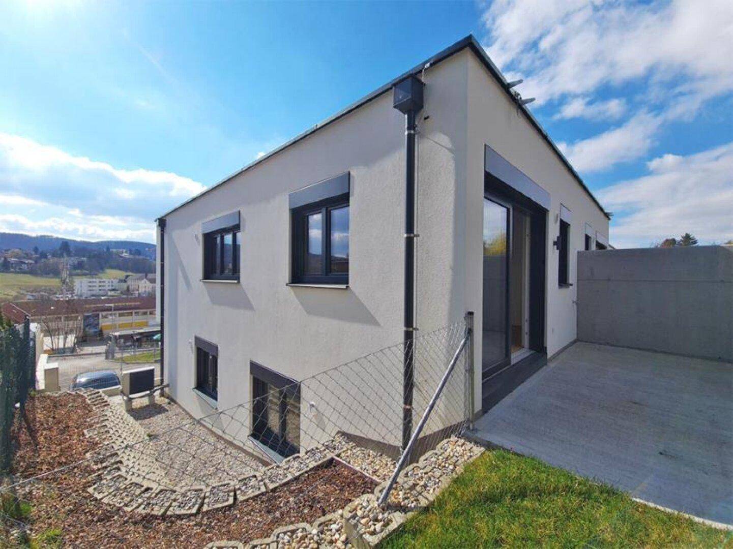 Moderne Hausfassade mit großen Fenstern und einer kleinen Betonfläche auf abschüssigem Gelände.