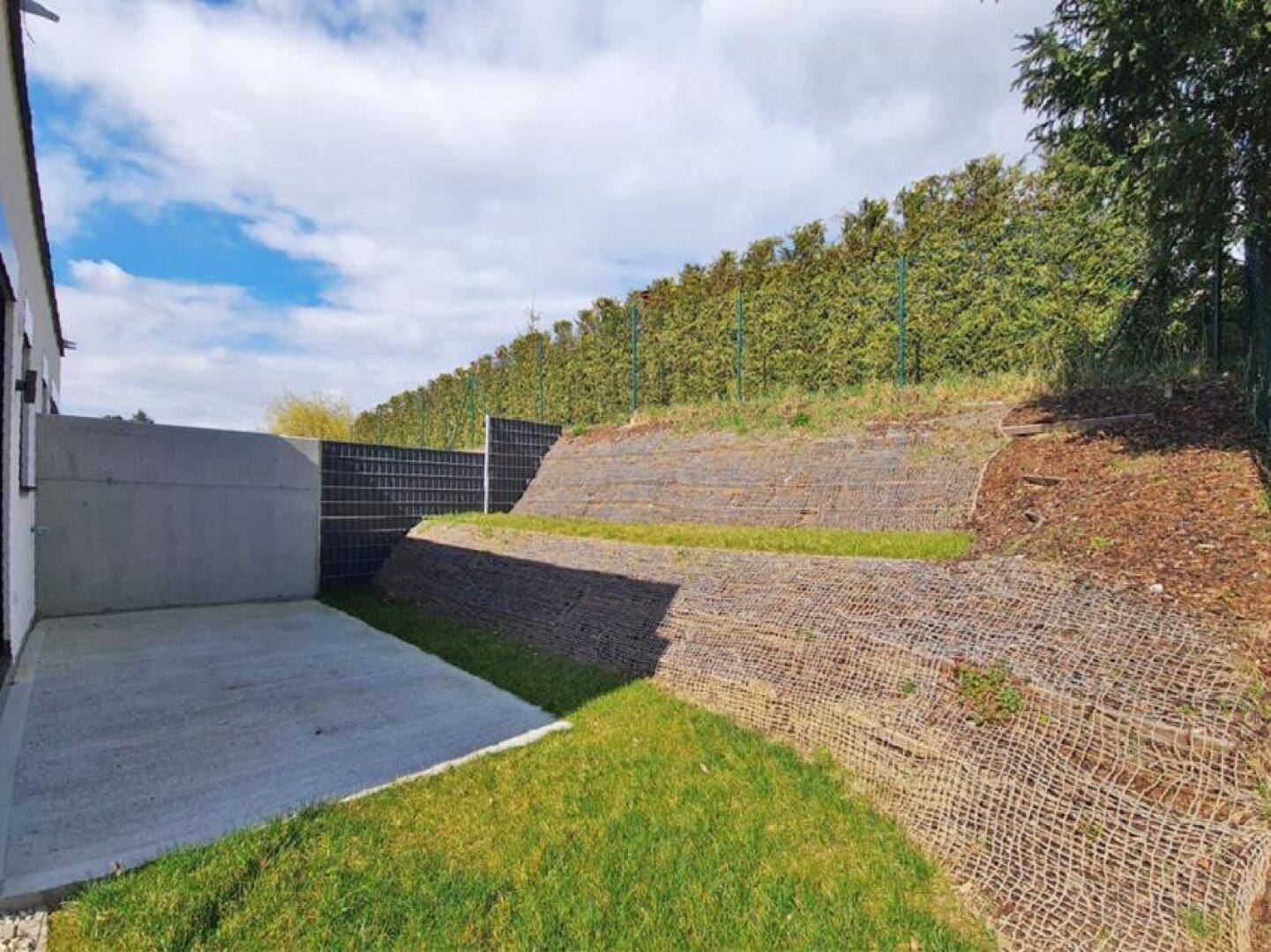 Terrassierter Gartenbereich mit Betonfläche und begrünten Böschungen, umgeben von Hecken.