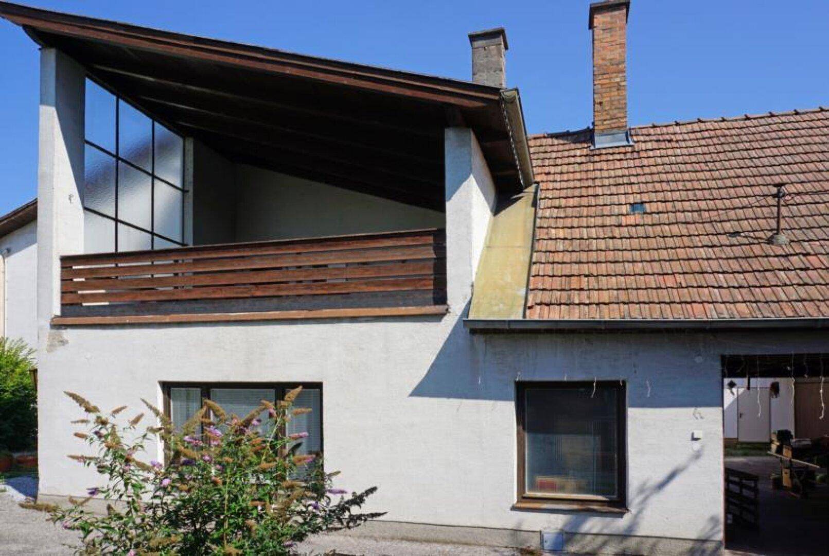 Überdachter Balkon mit Holzgeländer und großen Fenstern, angrenzend an ein älteres Gebäude.