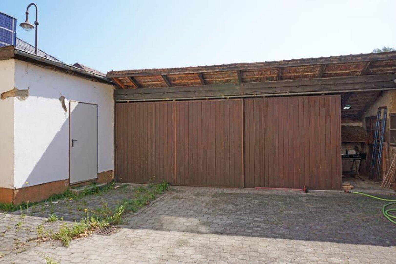 Großes Garagentor aus Holz und eine weiße Seitentür, mit gepflastertem Vorplatz.