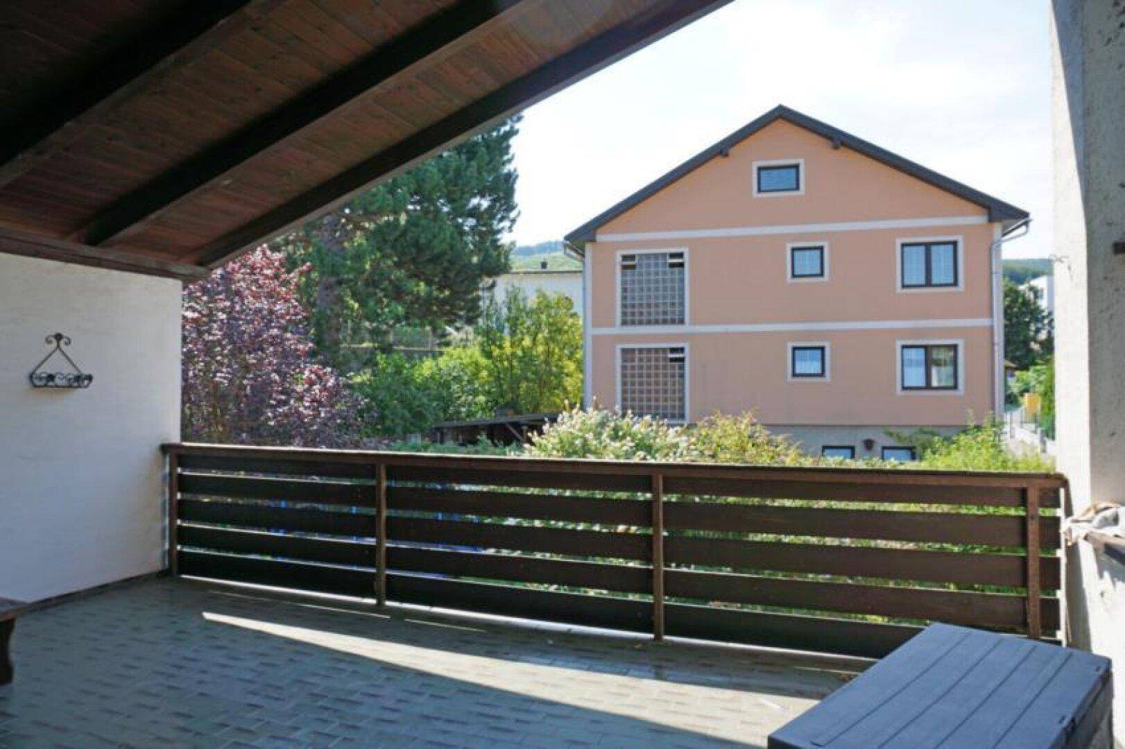 Überdachte Terrasse mit Holzgeländer und Blick auf ein benachbartes Gebäude und Bäume.