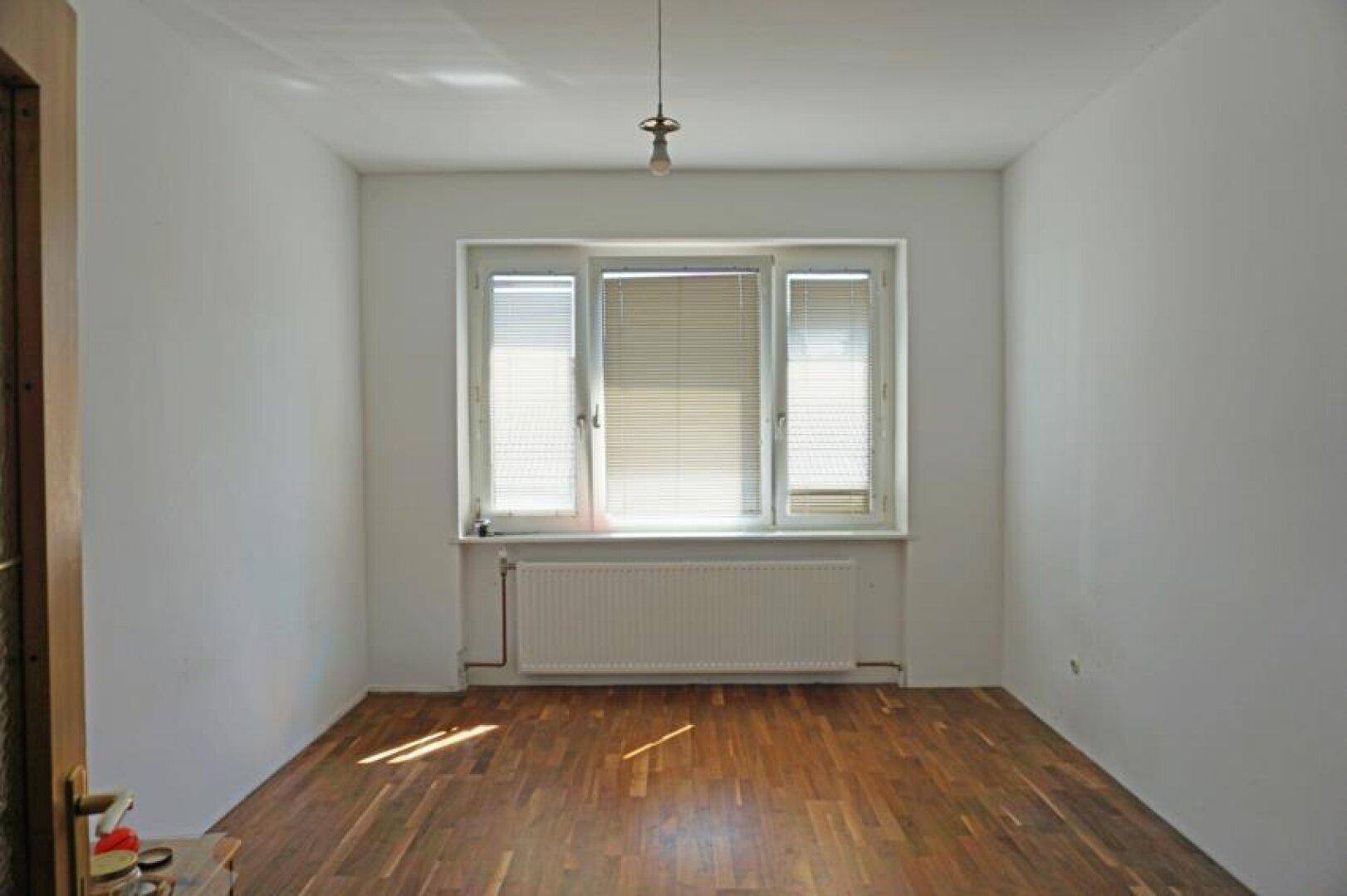 Leeres Zimmer mit Holzboden und Fenster, das viel Tageslicht hereinlässt.