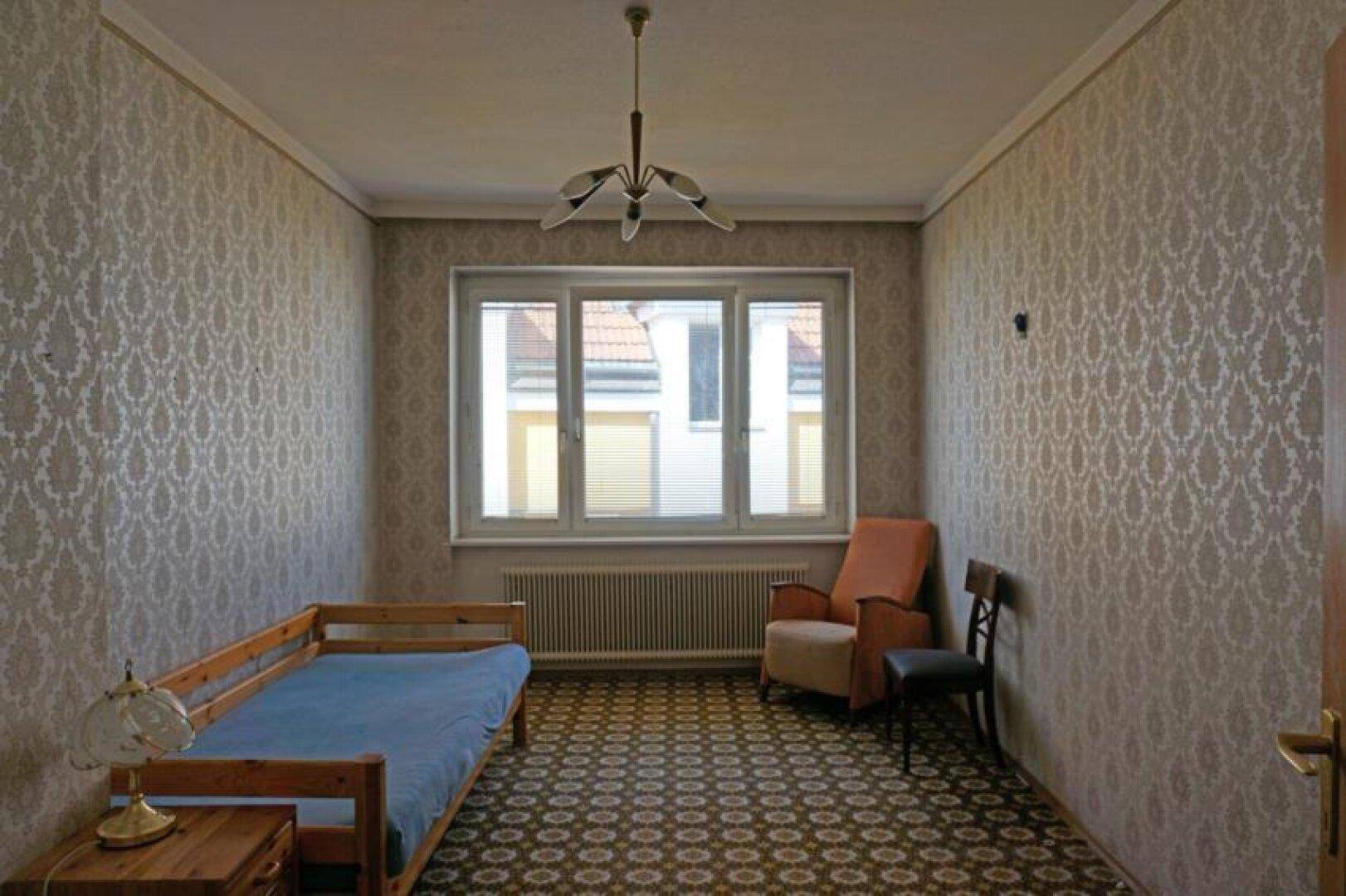 Schlafzimmer mit gemusterter Tapete, Teppichboden und einem Einzelbett, mit Blick nach draußen.