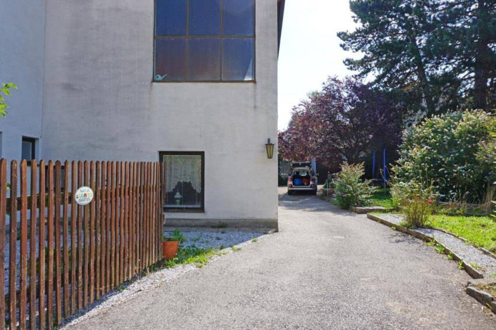 Schotterauffahrt neben einem Gebäude mit Holzzaun und Bäumen im Hintergrund.