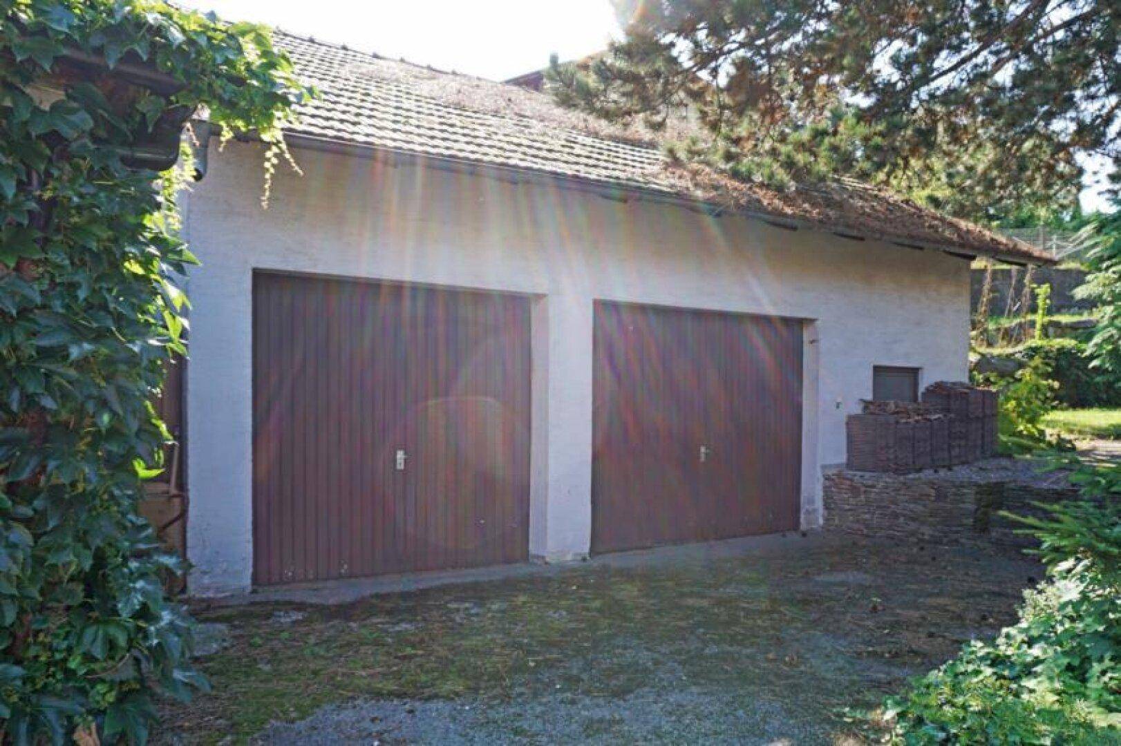 Doppelgarage mit Holztoren und umgebender Vegetation unter strahlendem Sonnenschein.