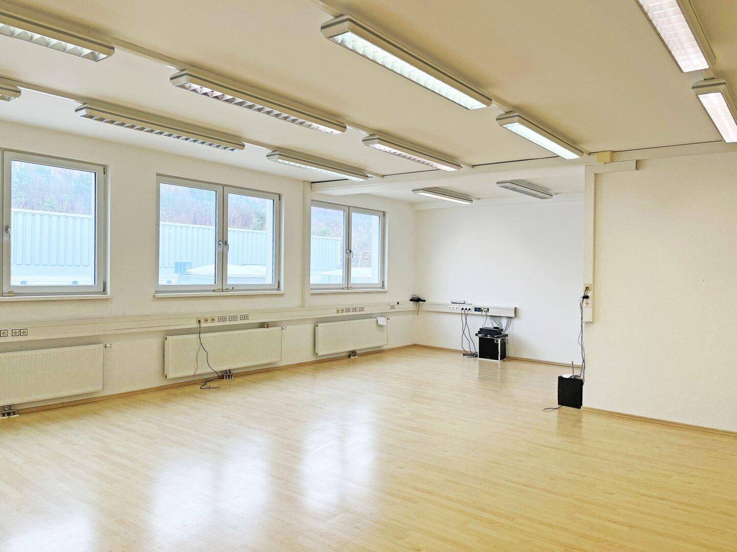 Geräumiger, heller Büroraum mit Holzfußboden, großen Fenstern und zahlreichen Deckenleuchten.