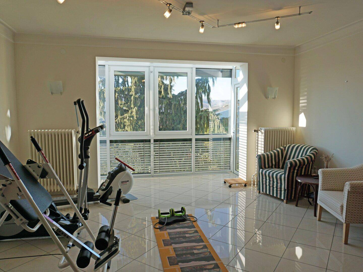 Heller Fitnessraum mit Trainingsgeräten, Fliesenboden und großen Fenstern.