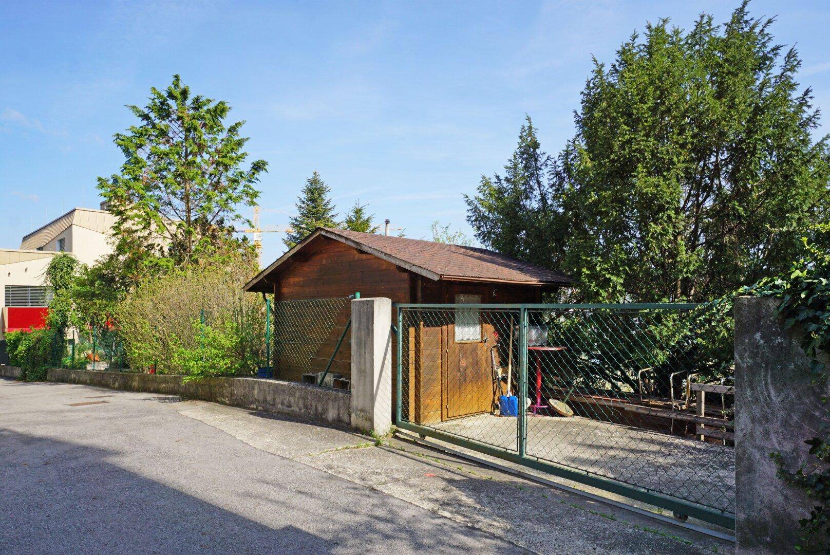 Gartenhaus aus Holz mit einem eingezäunten Bereich, umgeben von Bäumen und Grün.