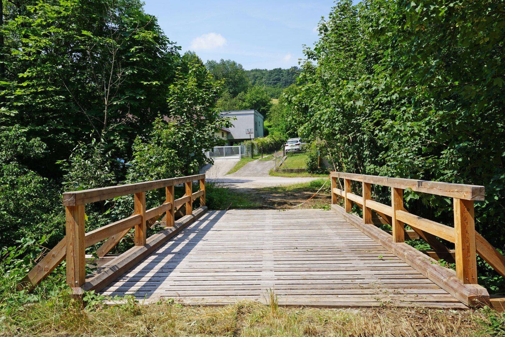 Hölzerne Brücke über einen Bach, die zu einem Grundstück mit einem Haus führt.