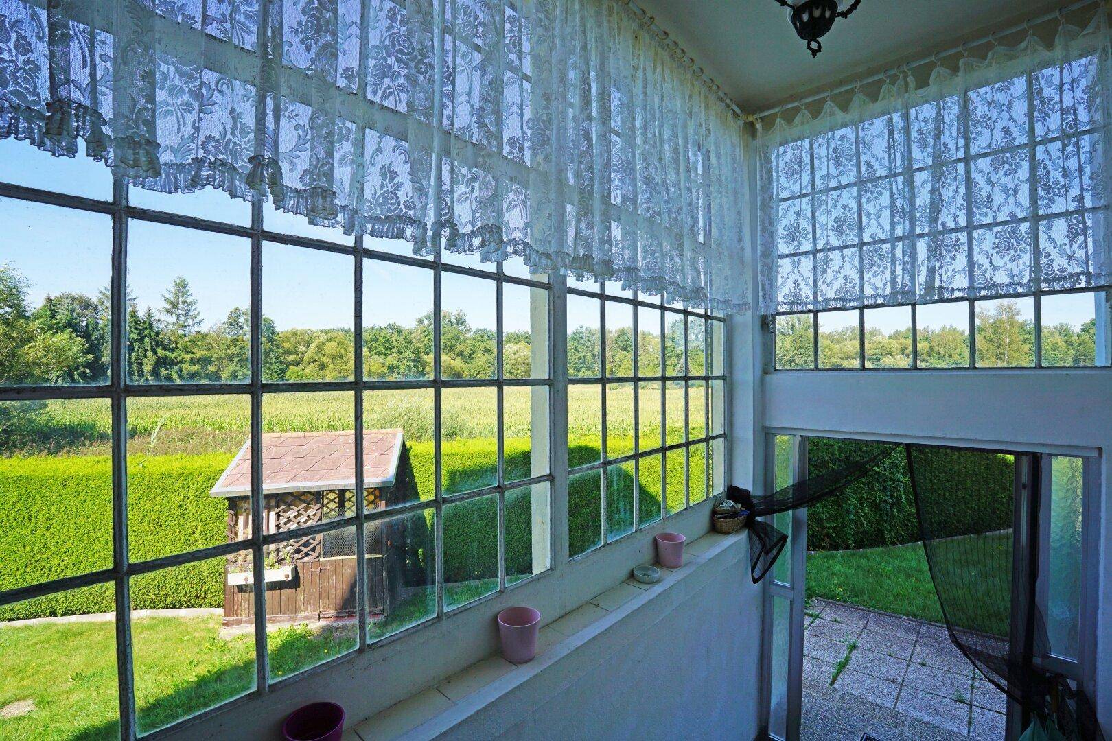 Verglaster Raum mit Blick auf den Garten, eine Hecke und die umliegende Natur.