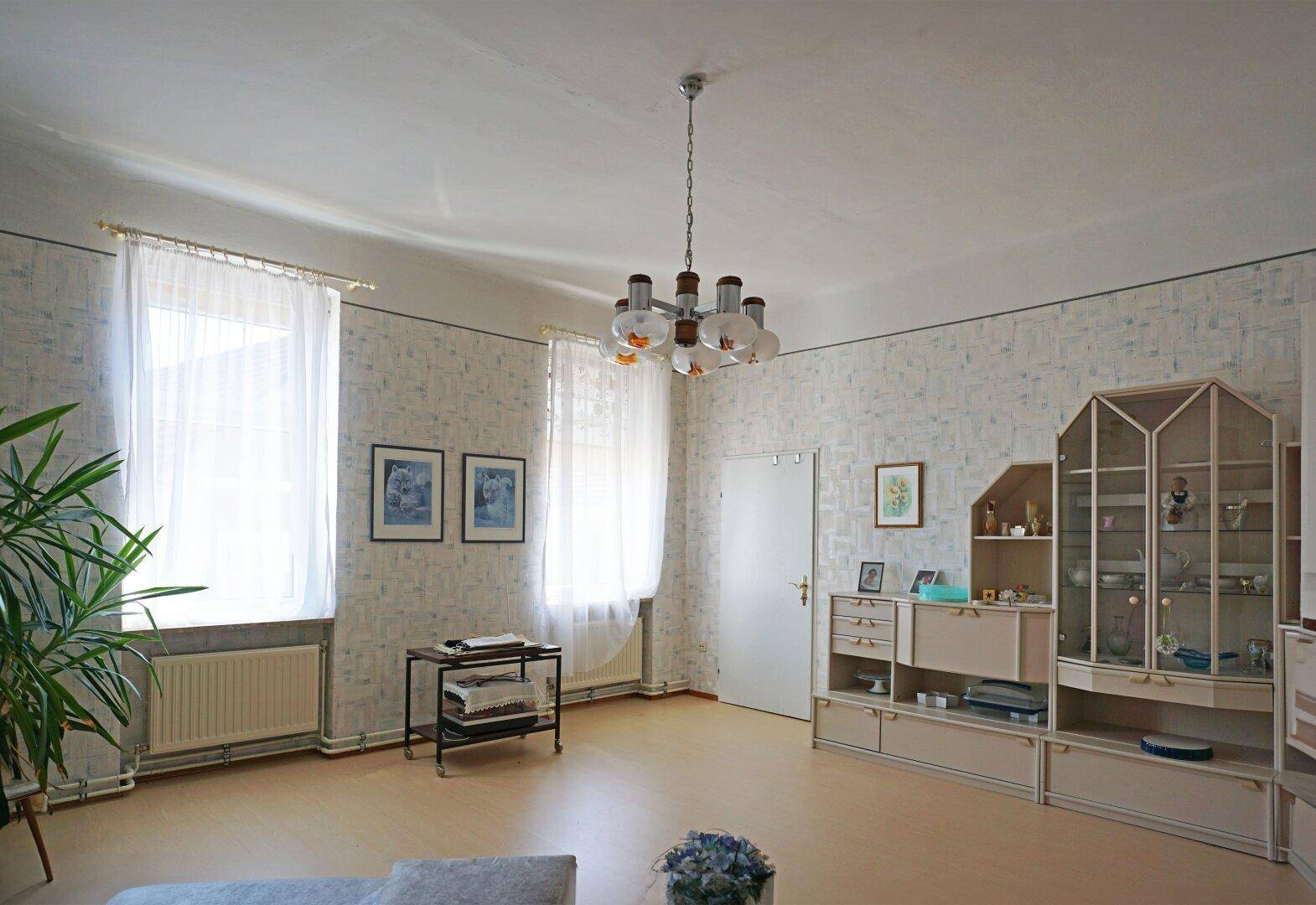 Wohnzimmer mit heller Tapete, Teppichboden und traditionellen Möbeln.