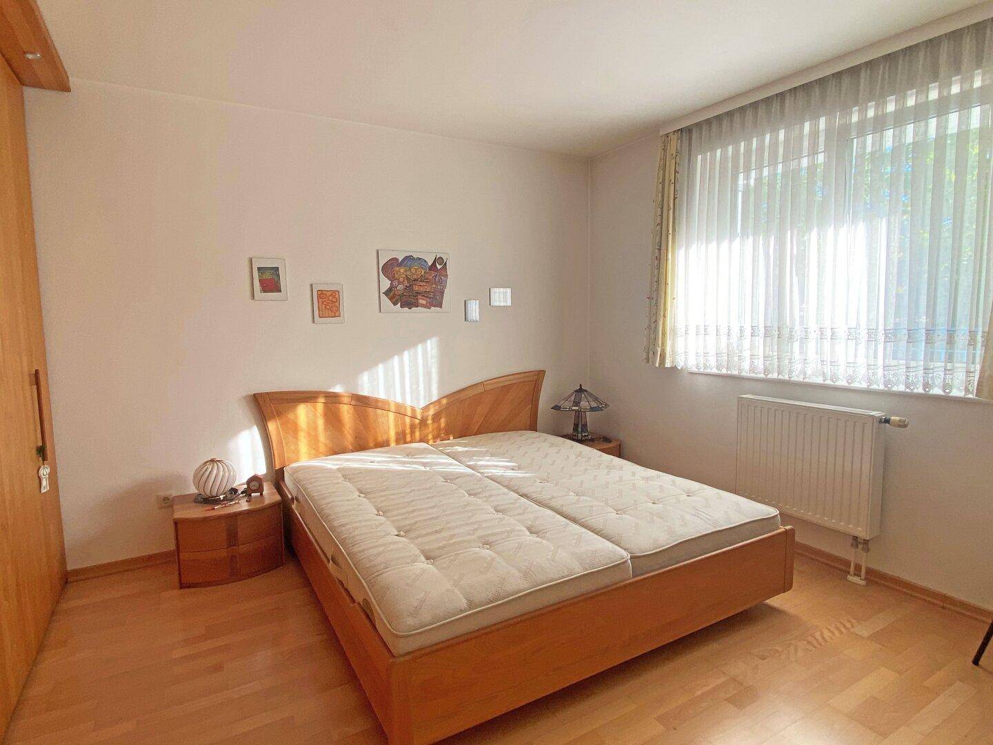 Helles Schlafzimmer mit Doppelbett, Holzmöbeln und Parkettboden unter sonnigem Fenster.