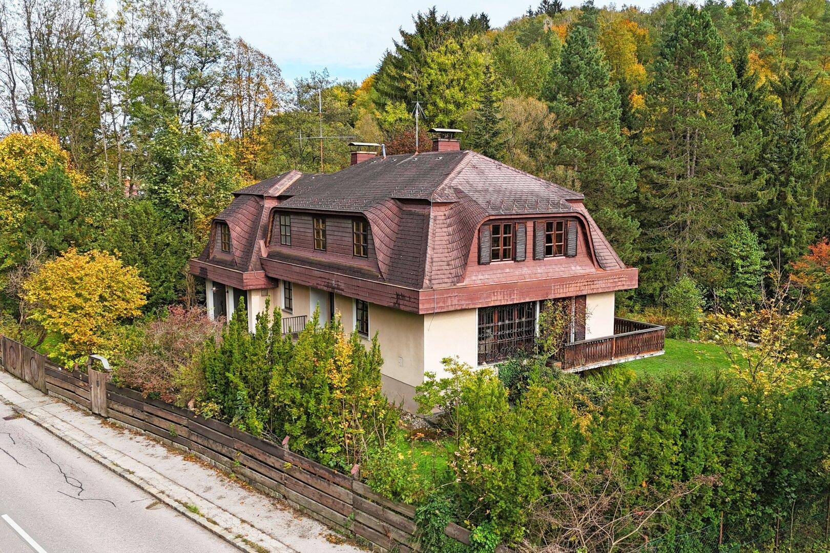 Ansicht des Hauses mit Mansarddach, umgeben von Bäumen und einem Zaun entlang der Straße.