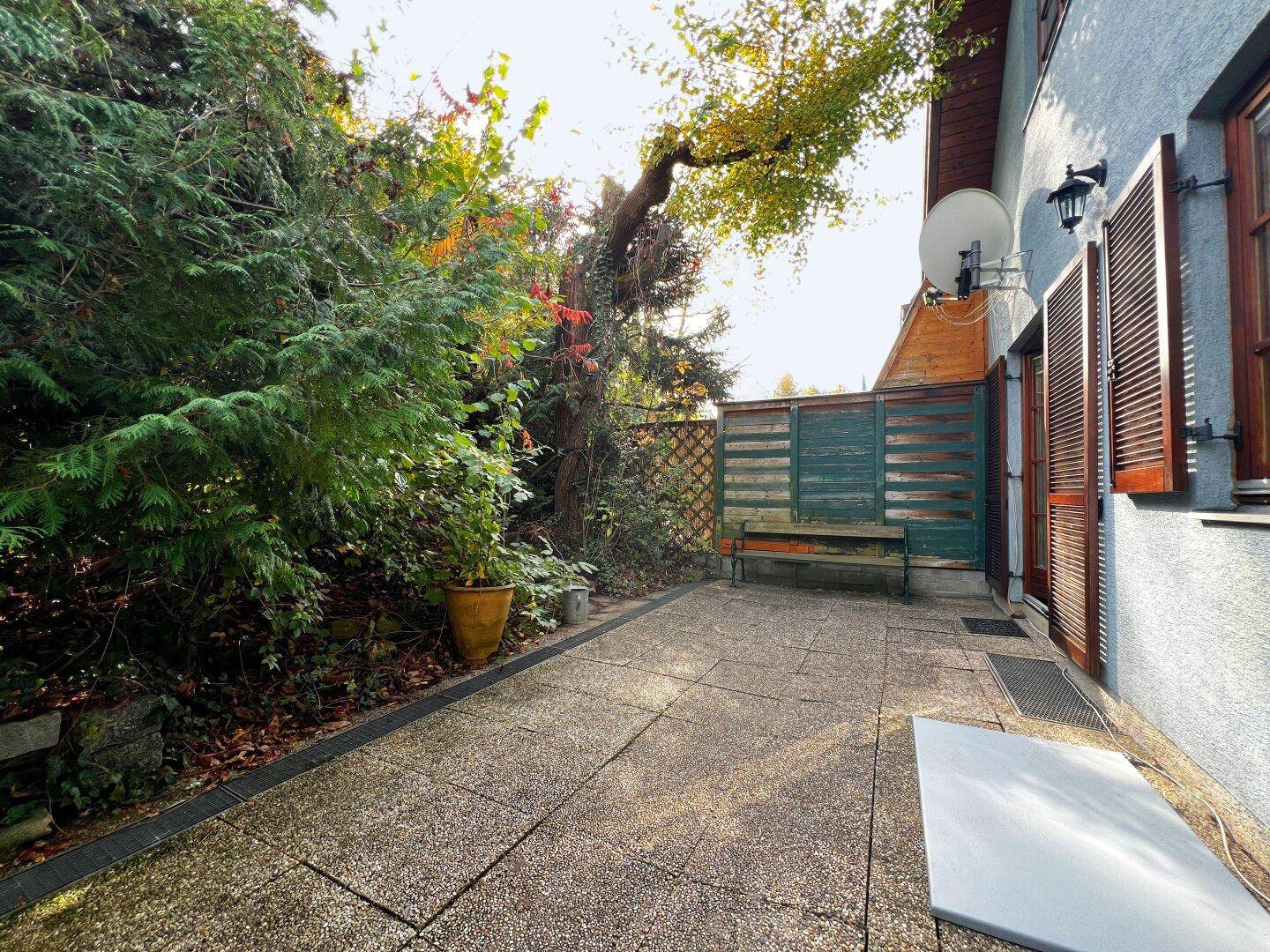 Gepflasterte Terrasse neben dem Haus mit einer Holzbank und üppigem Grün.