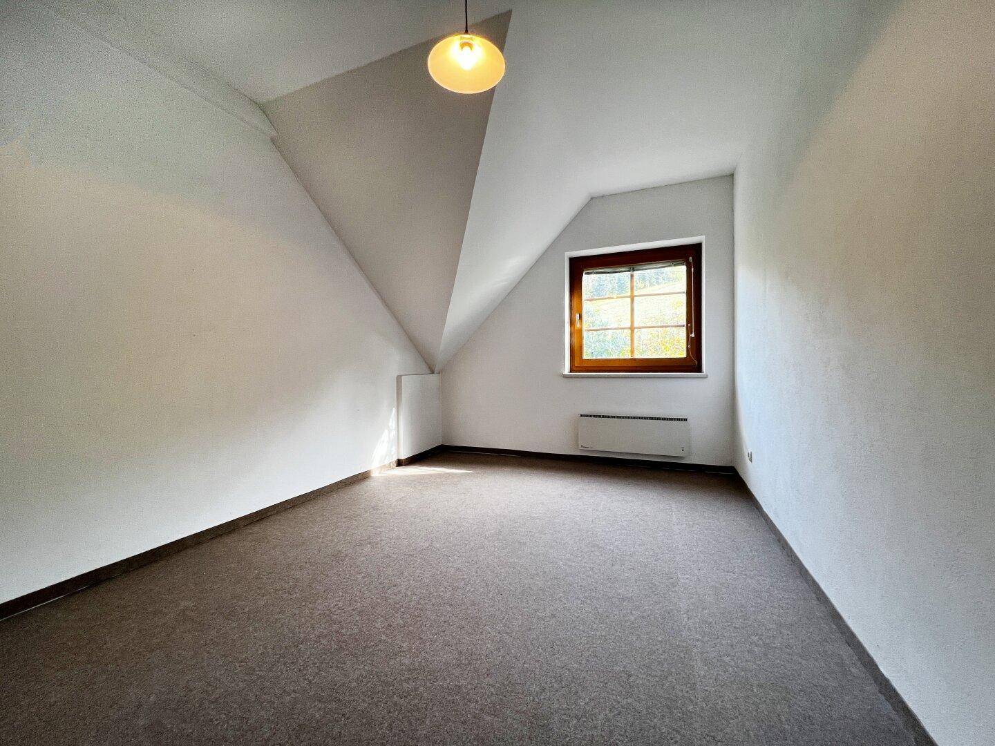 Schlafzimmer im Dachgeschoss mit schrägen Wänden, Teppichboden und einem Fenster.