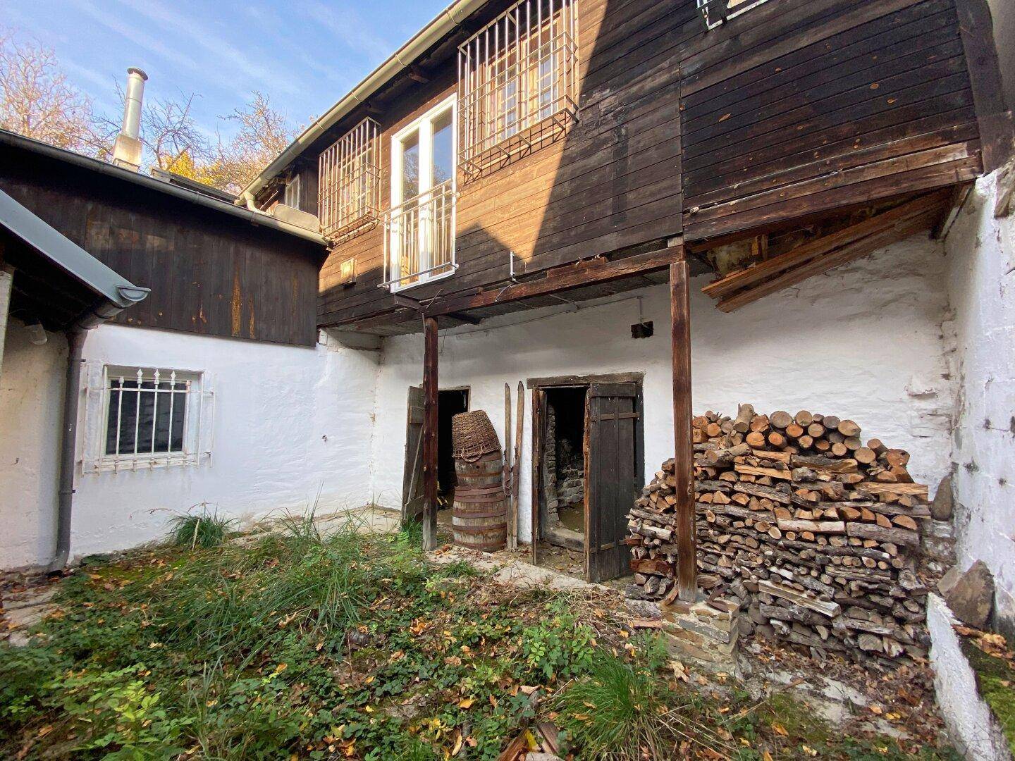 Hinterhofbereich mit gestapeltem Brennholz, weiß getünchten Wänden und einer oberen Holzfassade.