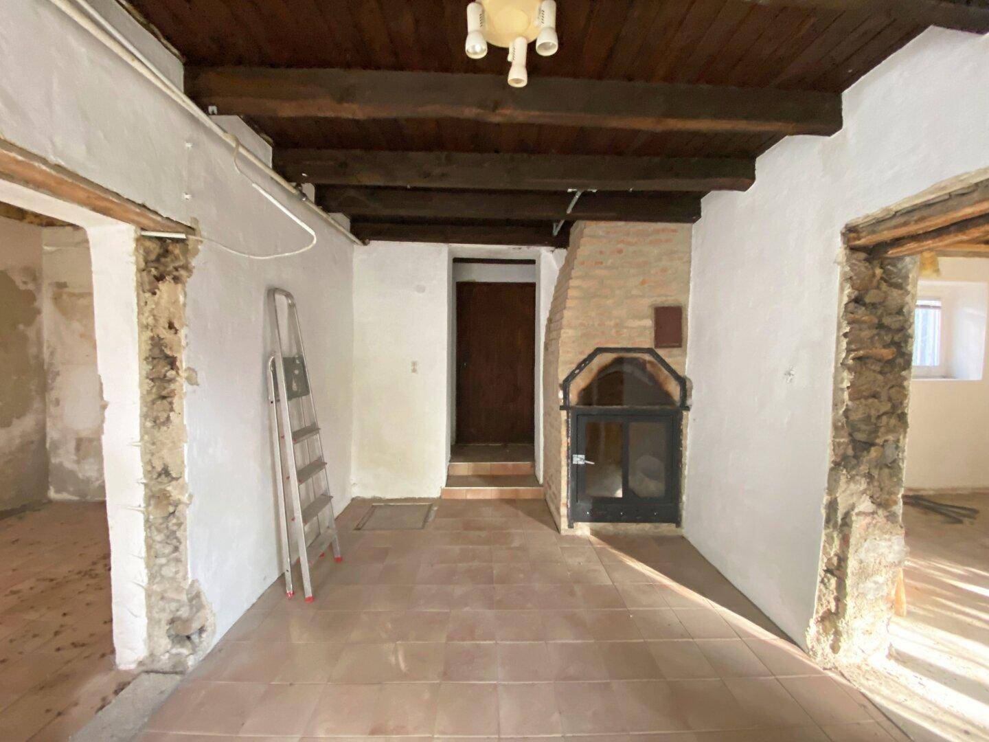 Innenraum in Renovierung mit sichtbaren Holzbalken, Fliesenboden und einem gemauerten Kamin.