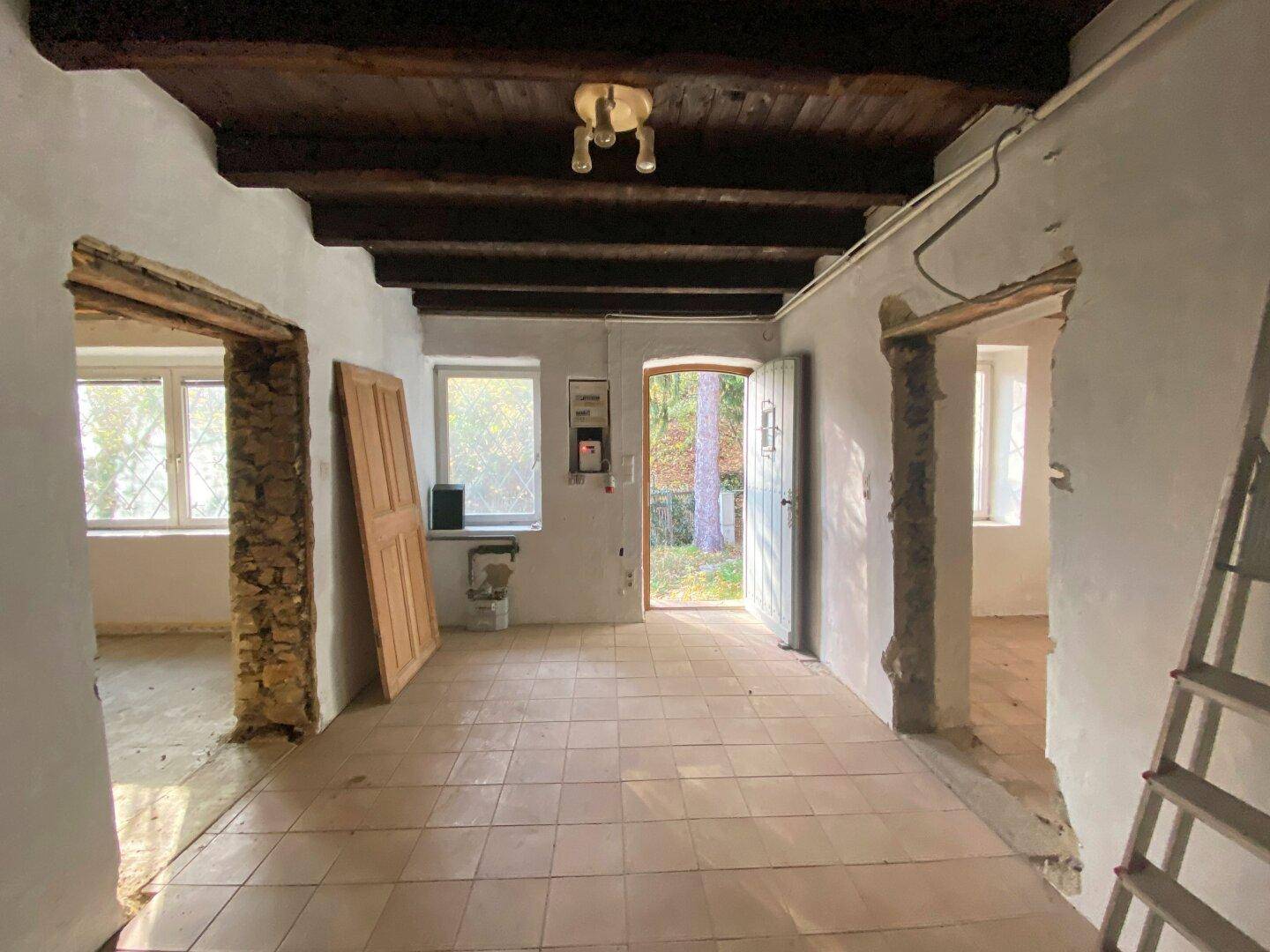 Renovierungsbedürftiger Raum mit Fliesenboden, Holzbalken und einem Ausgang ins Freie.