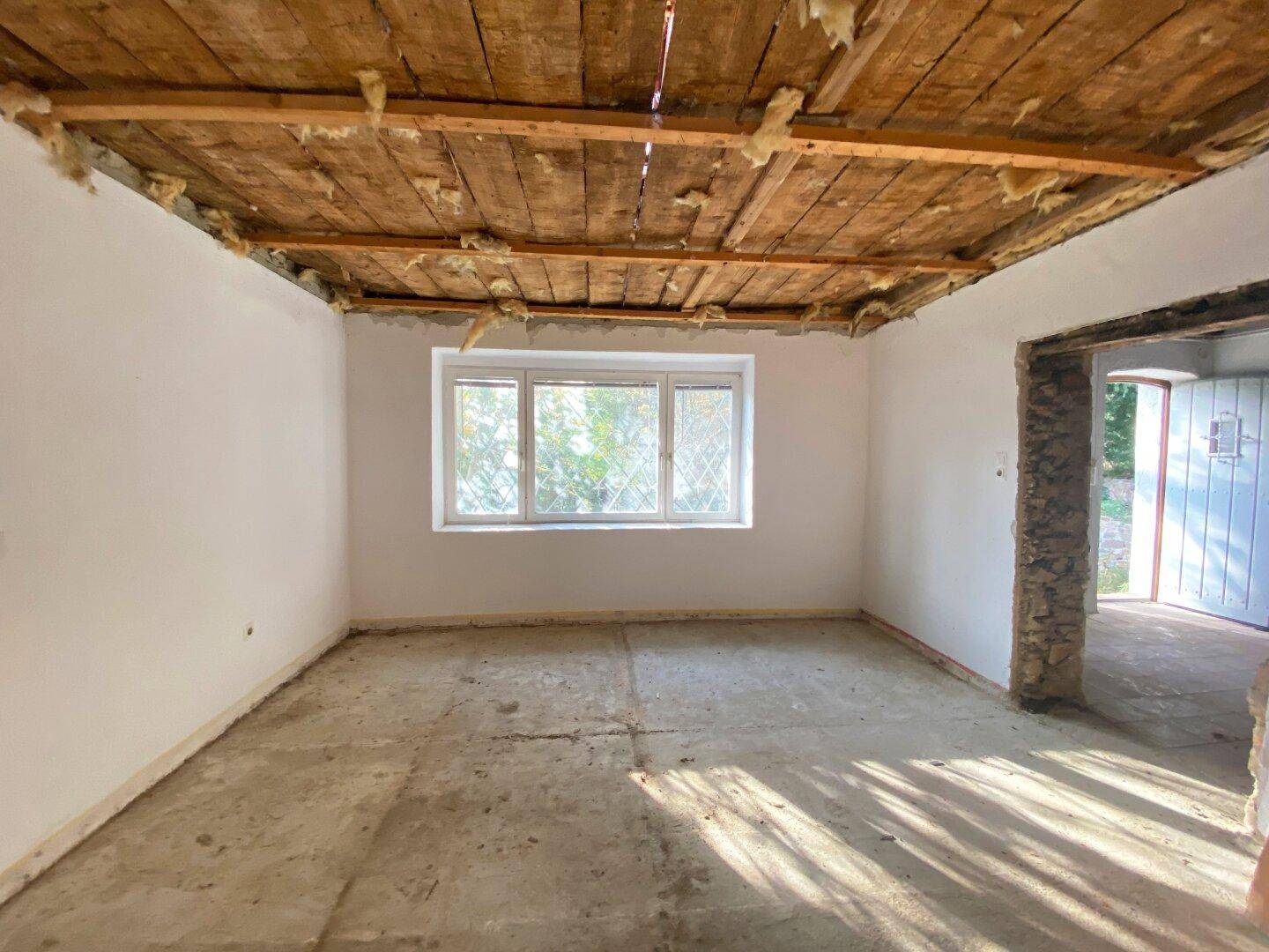 Renovierungsbedürftiger Raum mit Fenster, freiliegender Holzdecke und unfertigem Boden.