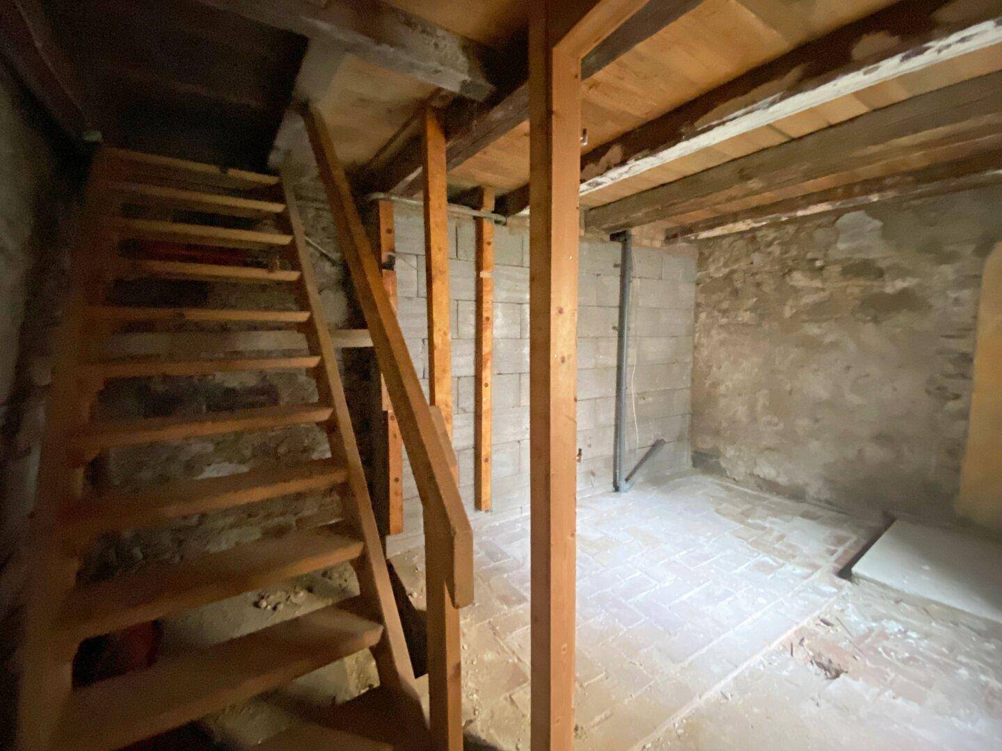Holztreppe führt hinunter in einen Kellerraum mit unverputzten Wänden und Fliesenboden.