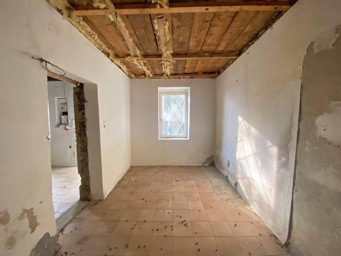 Renovierungsbedürftiger Raum mit Fliesenboden, Fenster und freiliegender Holzdeckenkonstruktion.