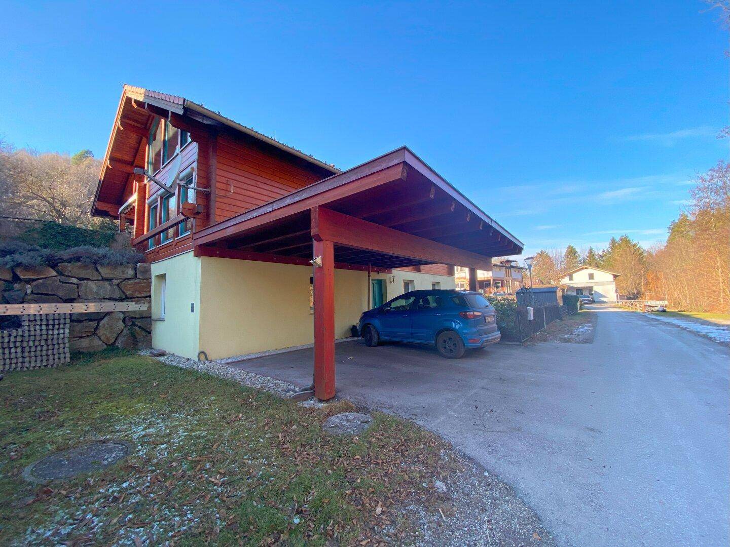 Ansicht des Holzhauses mit überdachtem Carport und Zufahrt an einem sonnigen Tag.