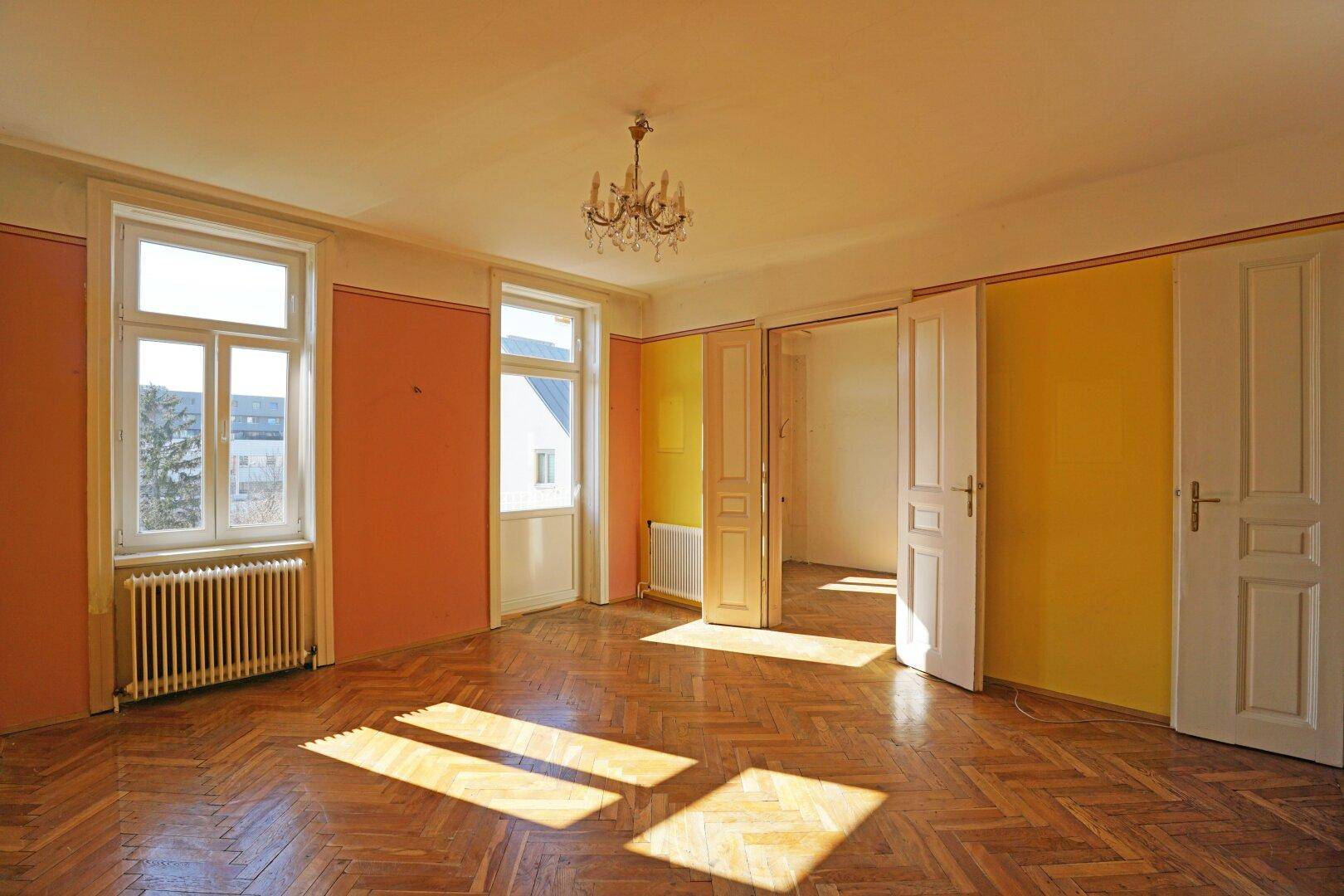Helles Wohnzimmer mit Fischgrätparkett, hohen Decken und großen Fenstern, die viel Tageslicht hereinlassen.