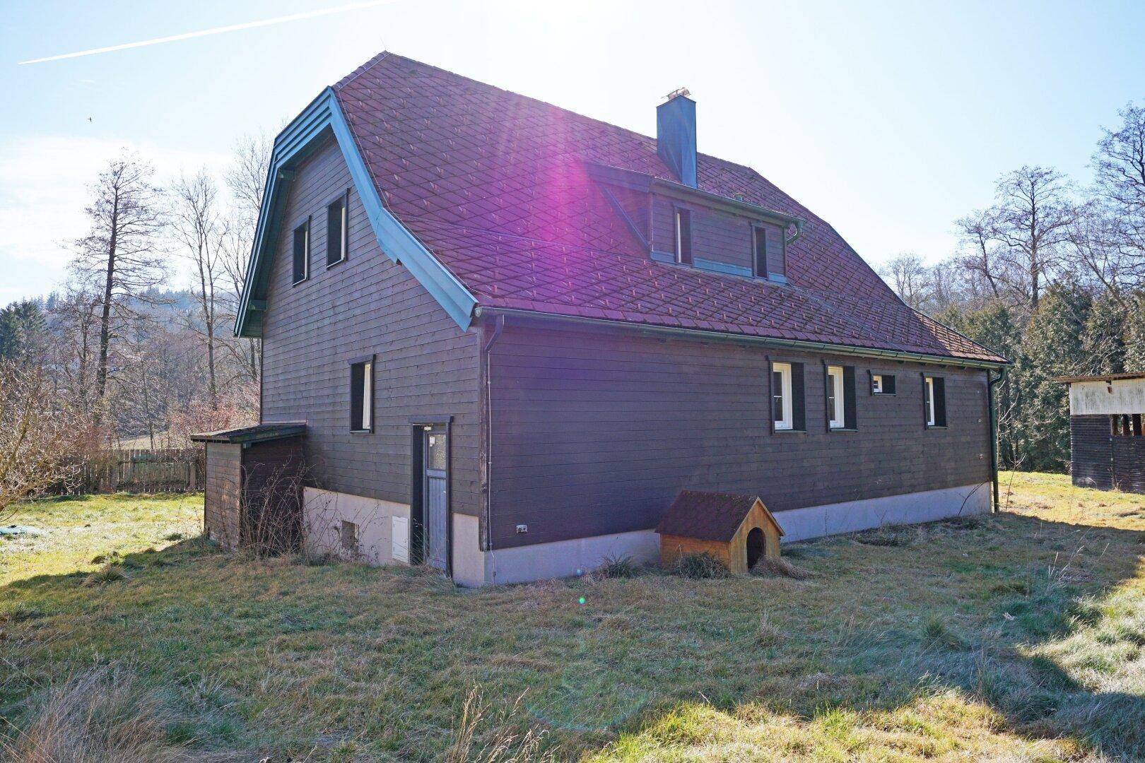 Rückansicht des Hauses mit Holzfassade, rotem Dach und einem kleinen Nebengebäude im weitläufigen Garten.