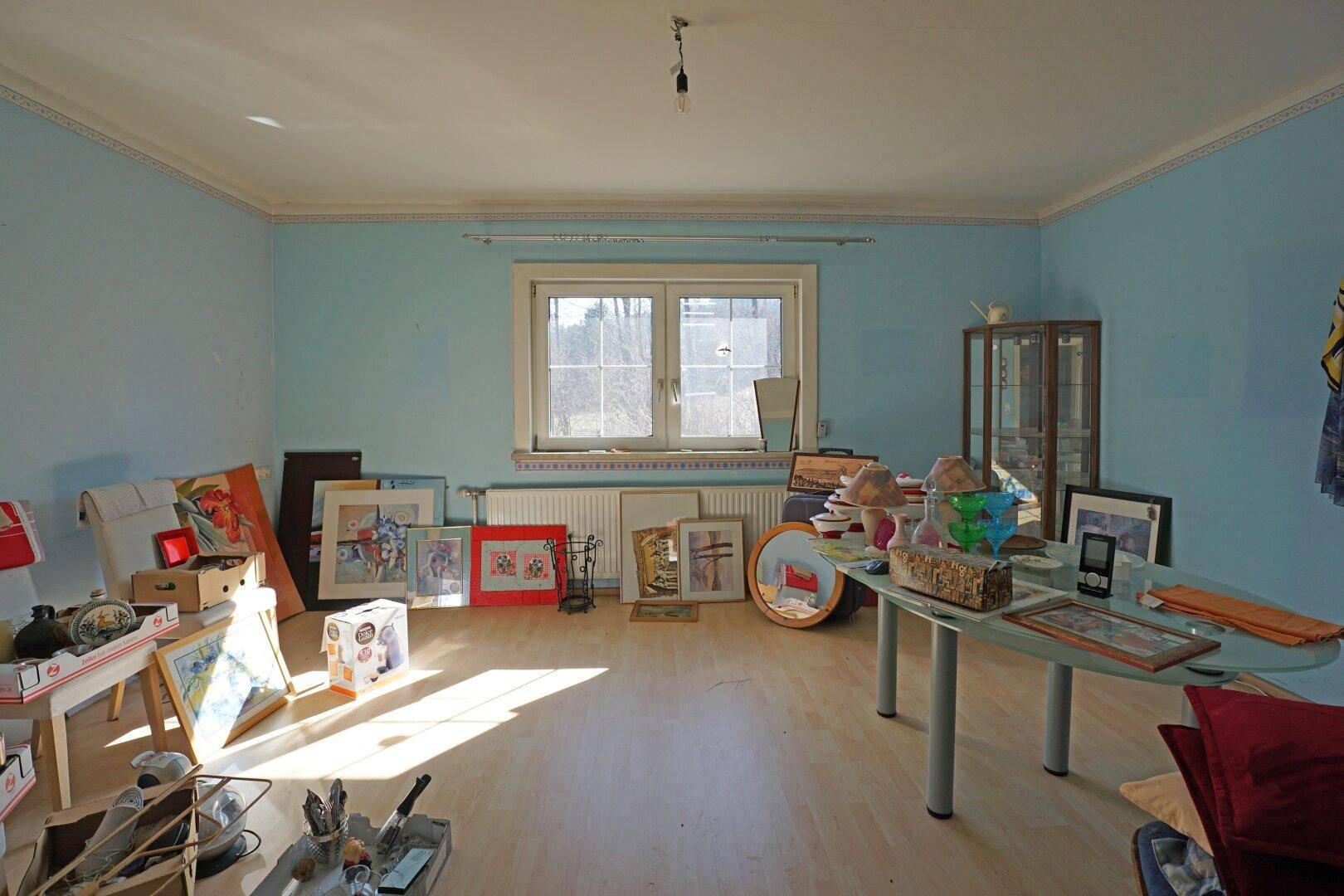 Helles Zimmer mit blauen Wänden, Stuckdecke und großem Fenster, derzeit als Abstellraum genutzt.
