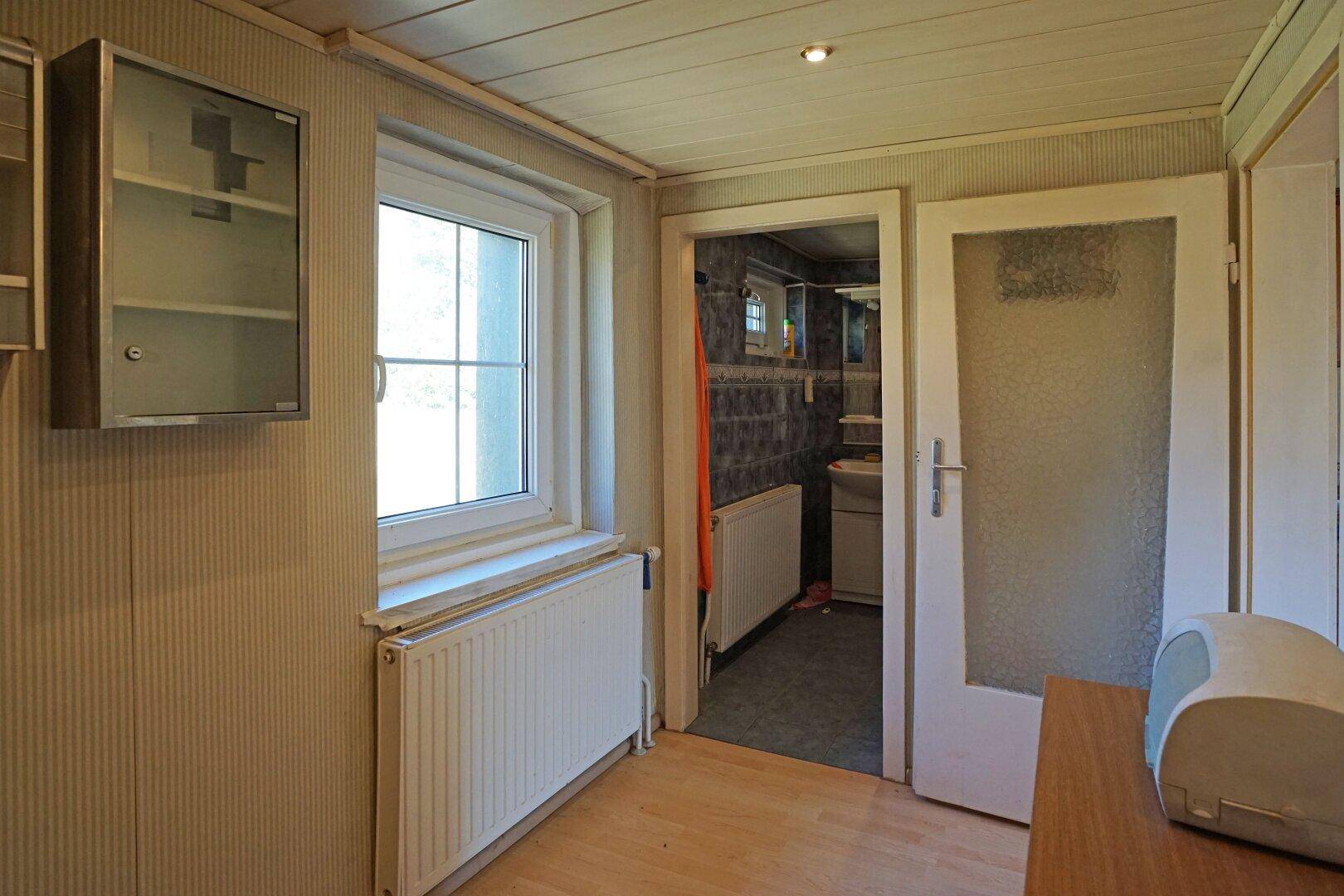 Schmaler Flur mit Fenster und Zugang zu einem Badezimmer mit dunklen Fliesen und Heizkörper.