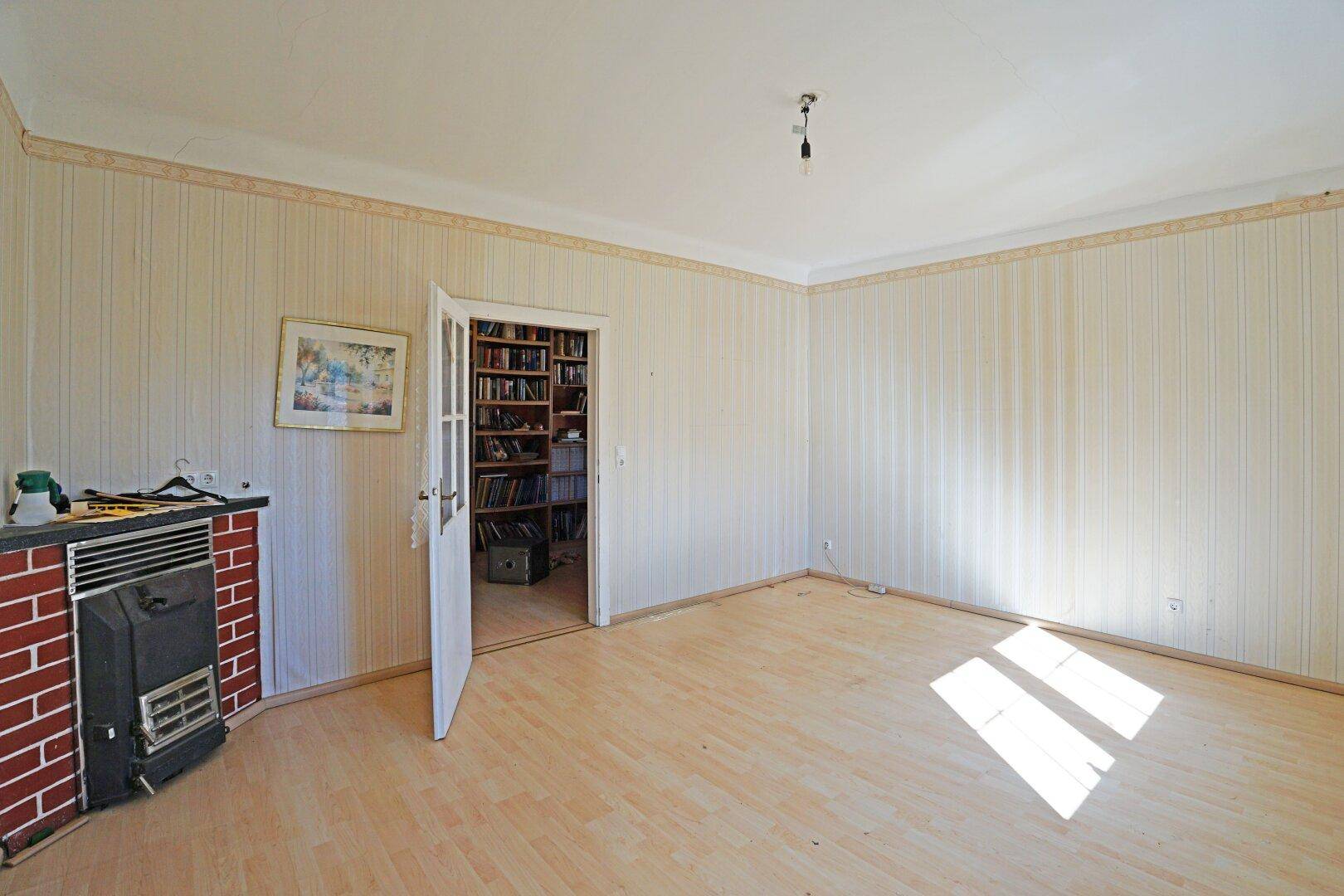 Geräumiges Wohnzimmer mit hellem Laminatboden, Kamin und Zugang zu einem weiteren Raum im Haus.