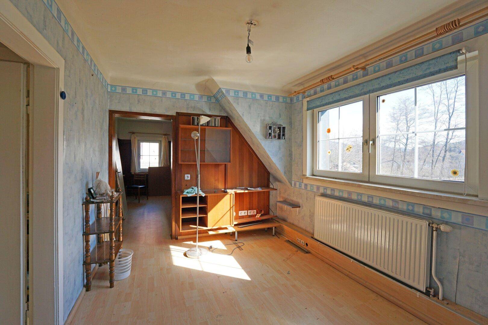 Helles Schlafzimmer mit Dachschräge, blauer Tapete und Blick ins Grüne durch das große Fenster.