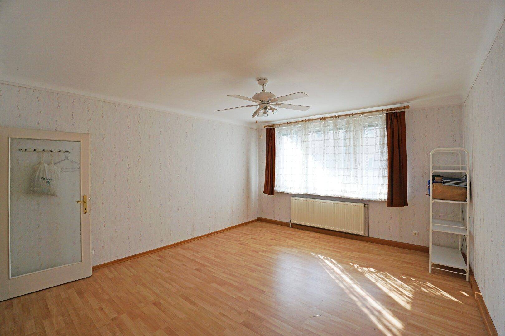 Helles, leeres Zimmer mit Laminatboden, Deckenventilator und großem Fenster, das viel Tageslicht hereinlässt.