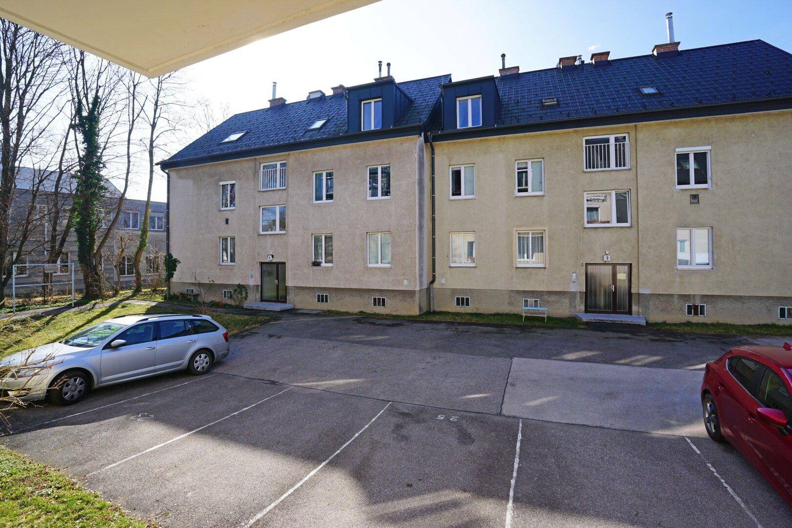 Mehrfamilienhaus mit Parkplätzen im Freien und Blick auf die umliegende Grünfläche.
