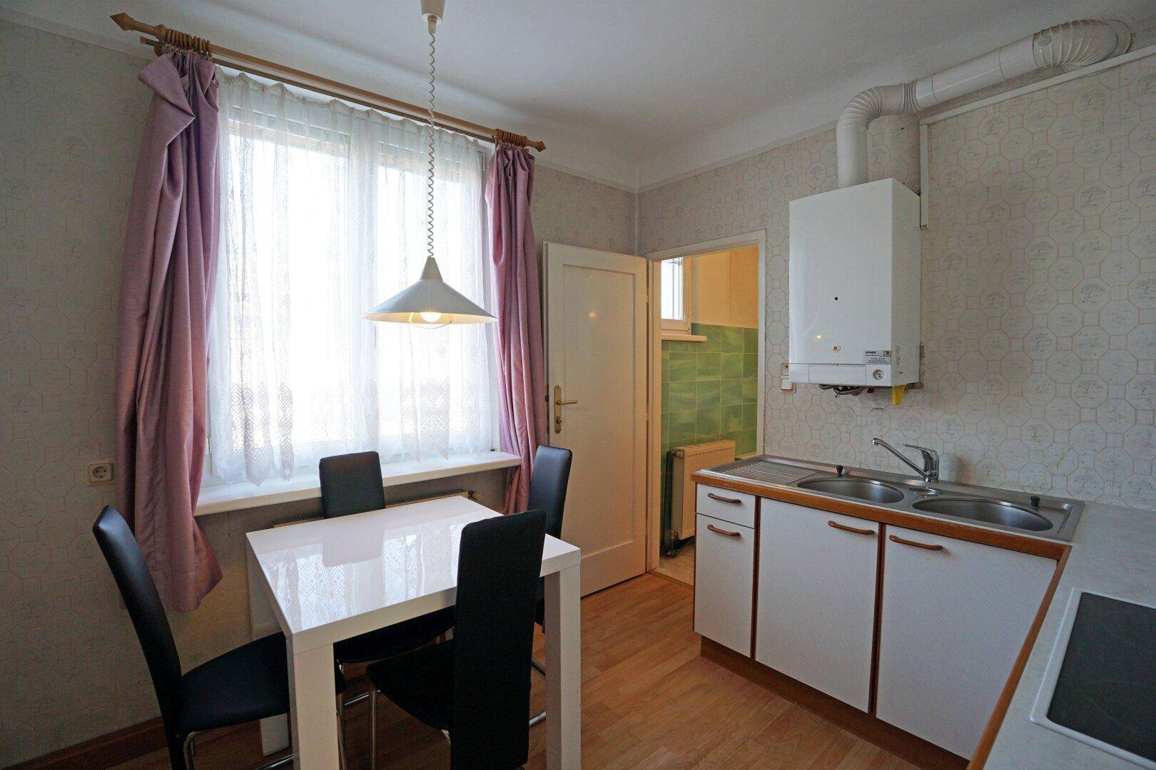Einfache Küche mit Spüle, Esstisch und Fenster, das Tageslicht hereinlässt.