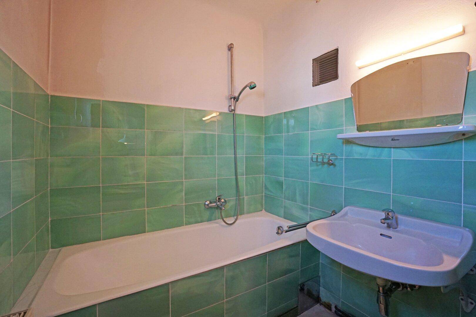 Badezimmer mit grünen Fliesen, Badewanne mit Duschfunktion und Waschbecken.