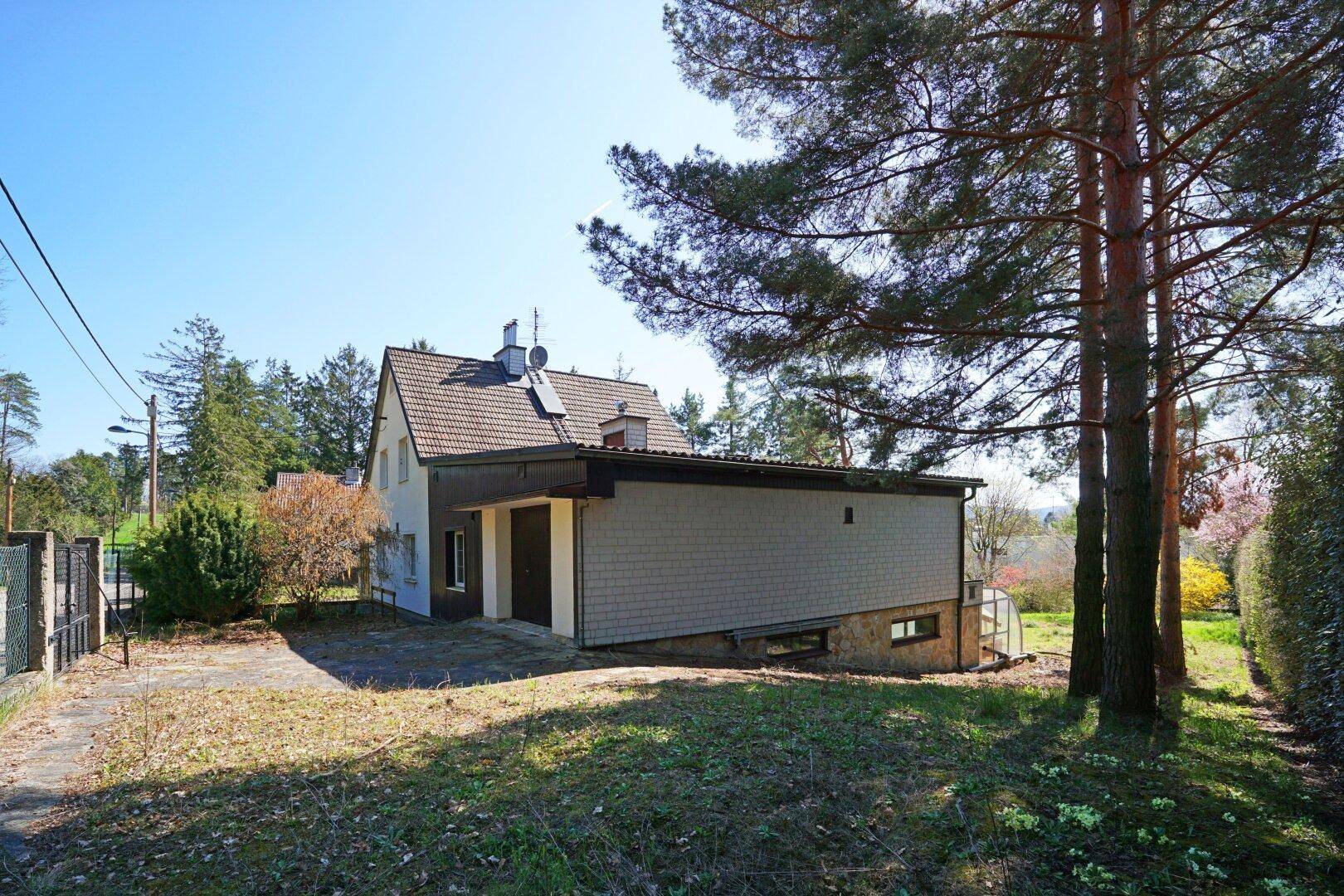 Seitenansicht des Hauses mit Anbau und Garage, umgeben von Bäumen und einer Rasenfläche.